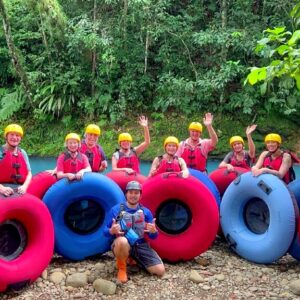 Large group of 11 adult travelers geared up for Rio Celeste tubing adventure, standing by the river's vibrant blue waters with their tour guide.