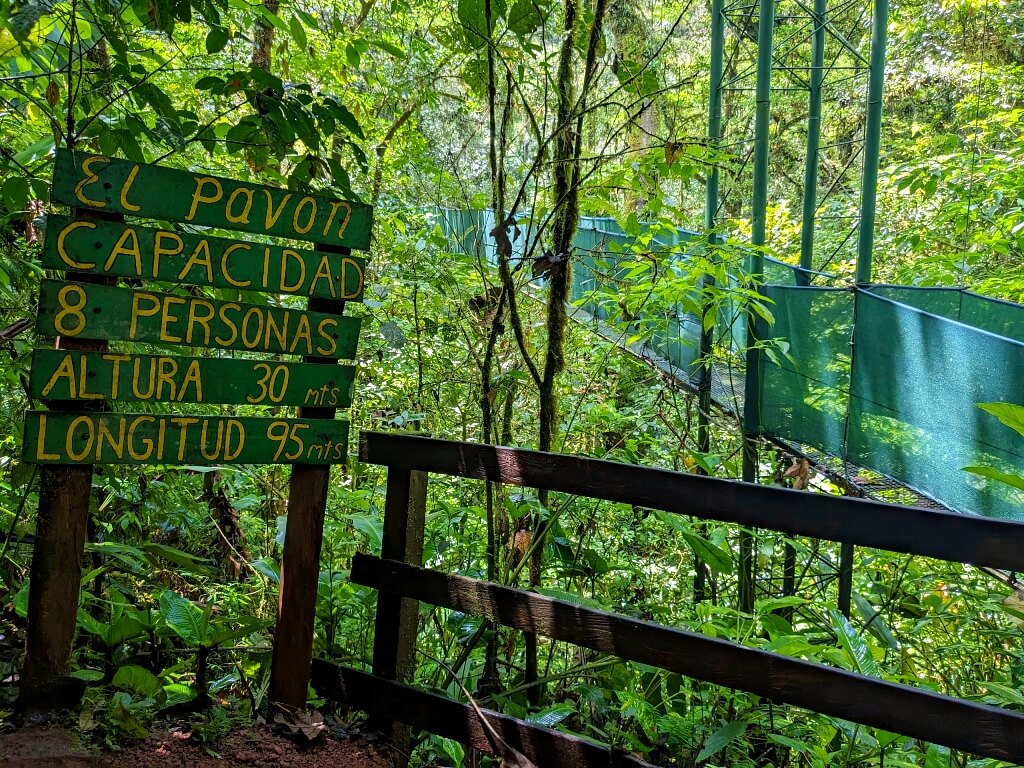 Sign post at the entrance of Hanging Bridge 'El Pavon' at Heliconias Private Reserve in Bijagua, displaying capacity for 8 people, height of 30 meters, and length of 95 meters.