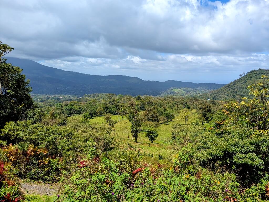Panoramic view of Bijagua valley showcasing lush greenery and diverse plant life, with a backdrop of the Miravalles Volcano mountain range and cloudy sky.