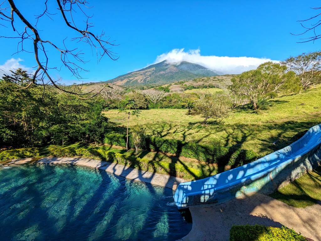Scenic view of blue water slide leading into a swimming pool, with pastures, trees, and Miravalles Volcano in the background at hot springs near Bijagua