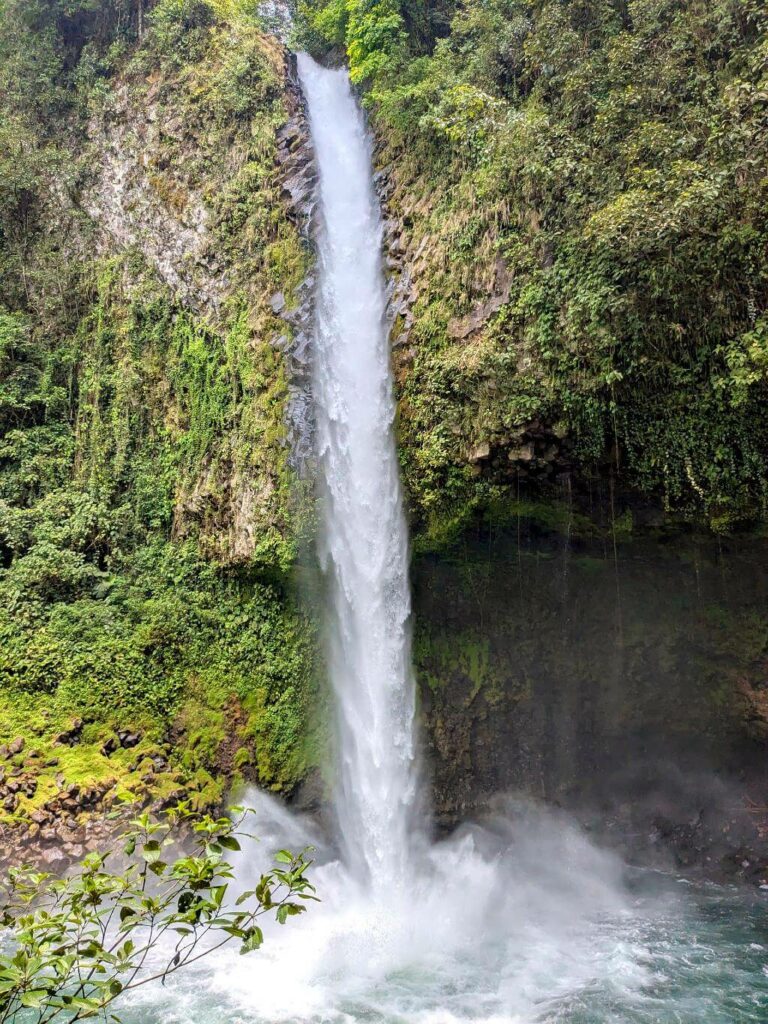 Close-up of La Fortuna Waterfall, showing white water crashing into the pool and mist rising, with the cliff and lush forest above.