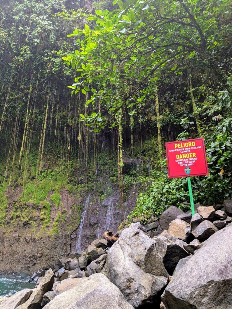 Lush vegetation and danger sign at the base of La Fortuna Waterfall, warning visitors to stay within authorized areas.