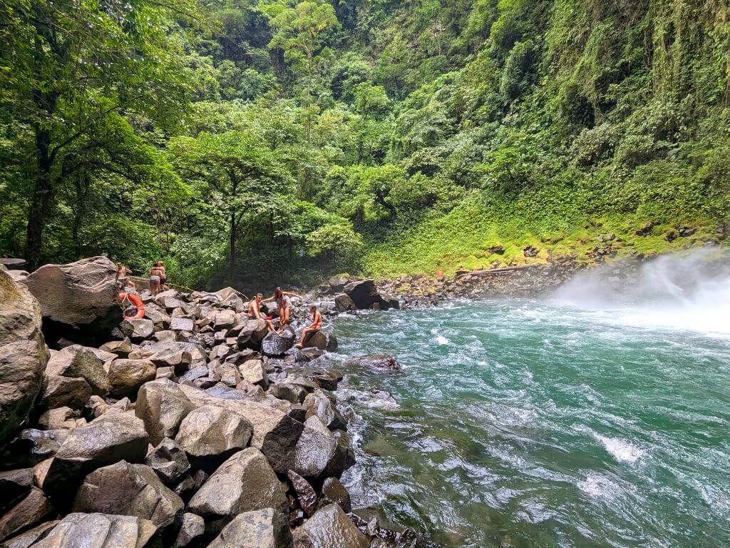 People resting on rocks around the pool at the base of La Fortuna Waterfall, with mist and rainforest in the background.