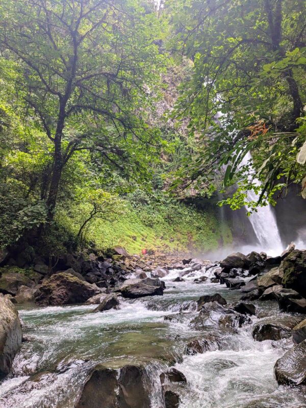 View of La Fortuna Waterfall from downstream, highlighting the clear waters, volcanic rocks, and lush forest.