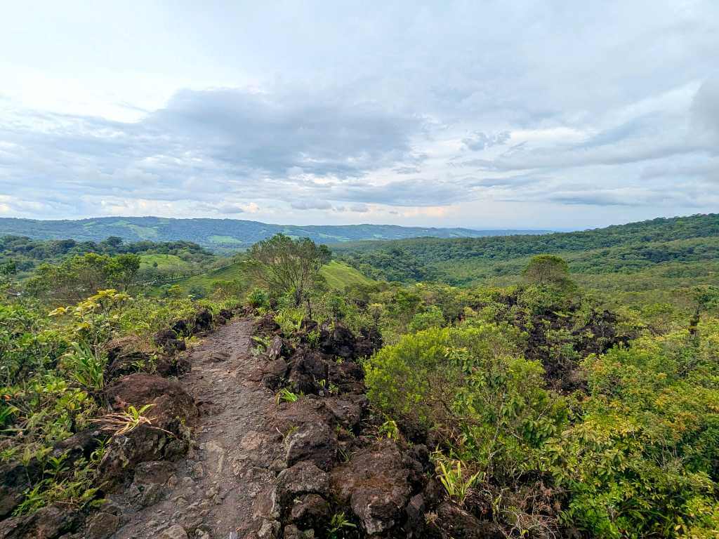 Lava flow trail at Arenal Volcano sprinkled with new plant growth, with views of the rainforest and mountains in the background.