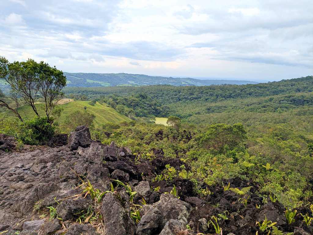 Panoramic view from Arenal Volcano's foothills featuring distant mountains, a green lagoon, and a landscape marked by solidified lava flows and lush rainforests.