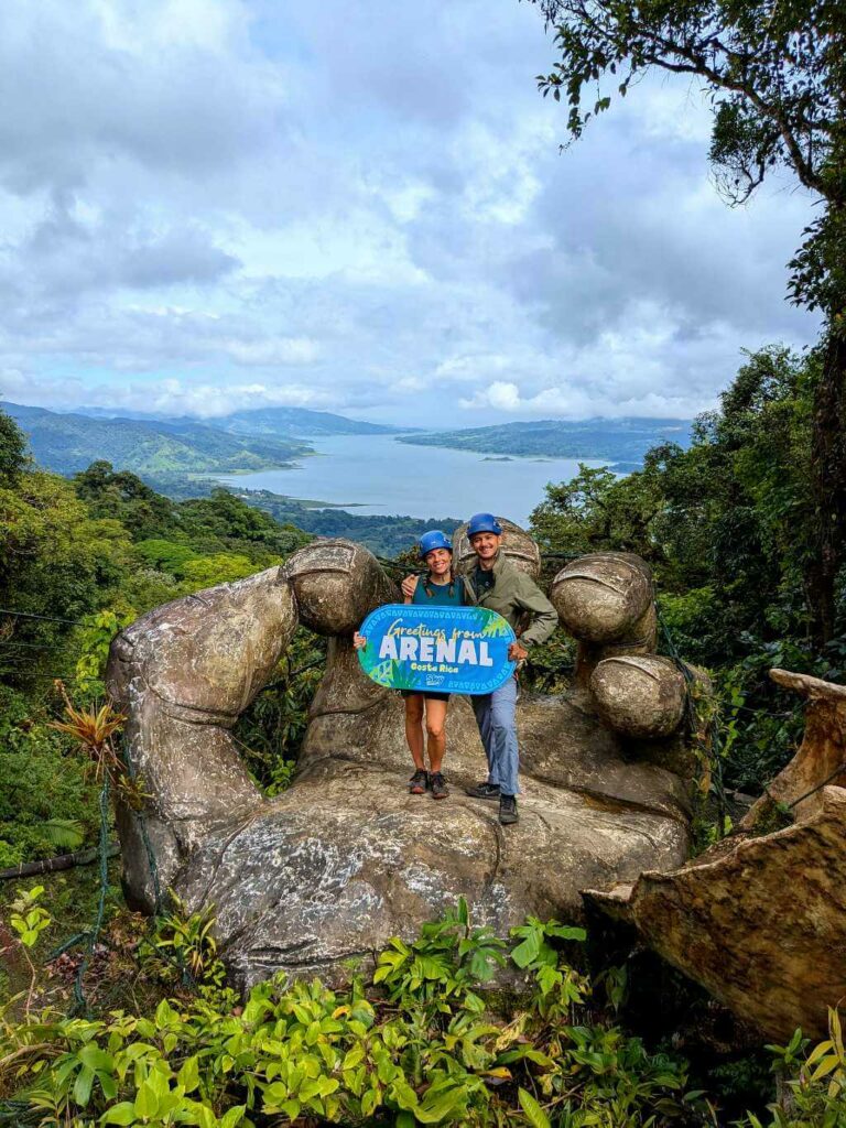 Travelers posing on the "Hand of Arenal" sculpture with Arenal Lake in the background.