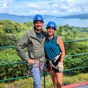 Two travelers smiling with Arenal Lake and lush rainforest in the background.