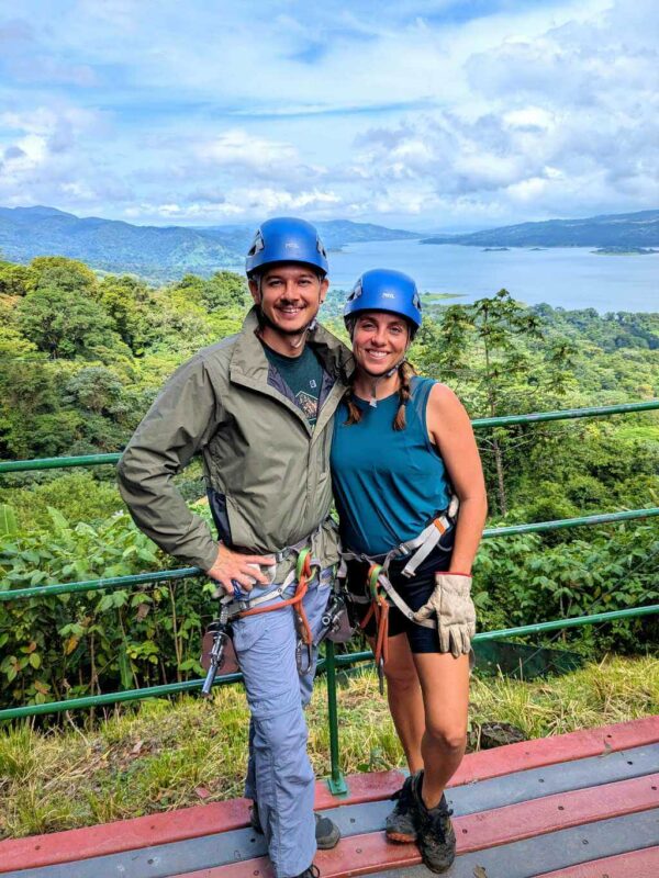 Two travelers smiling with Arenal Lake and lush rainforest in the background.