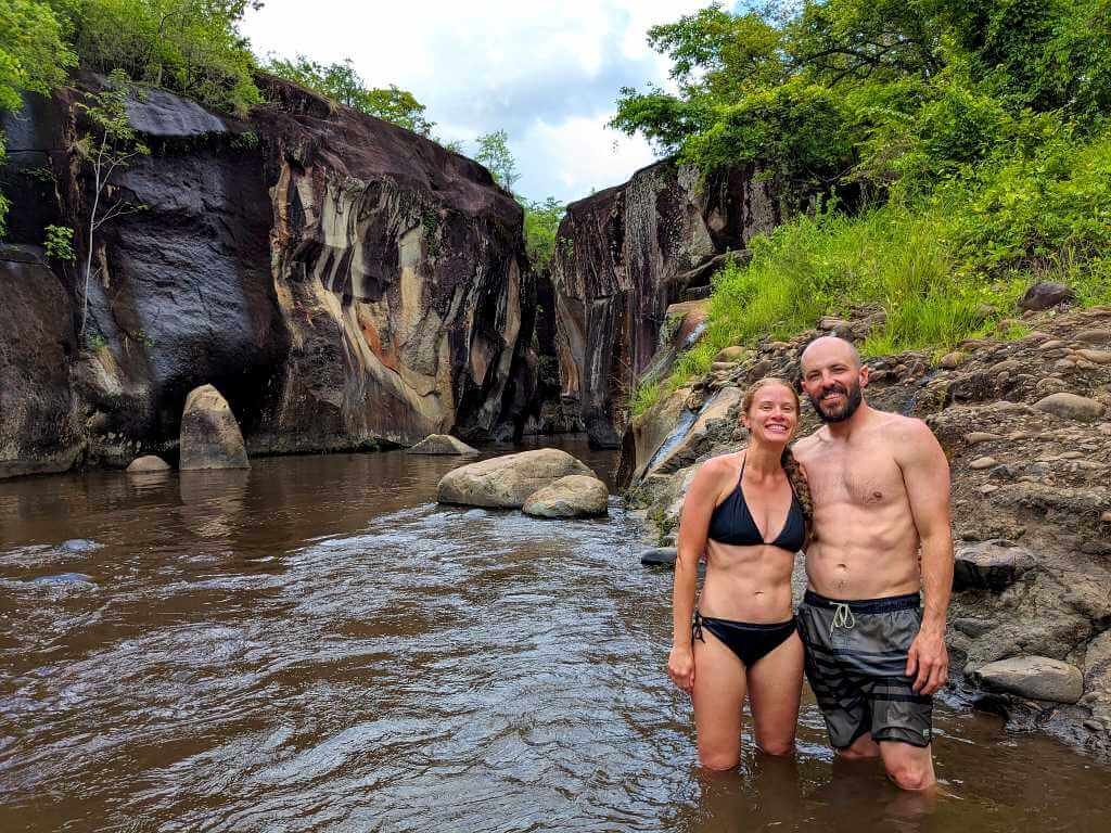 Two travelers pose at Pozas del Cañón in Rio Colorado after jumping from canyon walls into the deep river pool, with ancient petroglyphs visible on the rock surfaces.