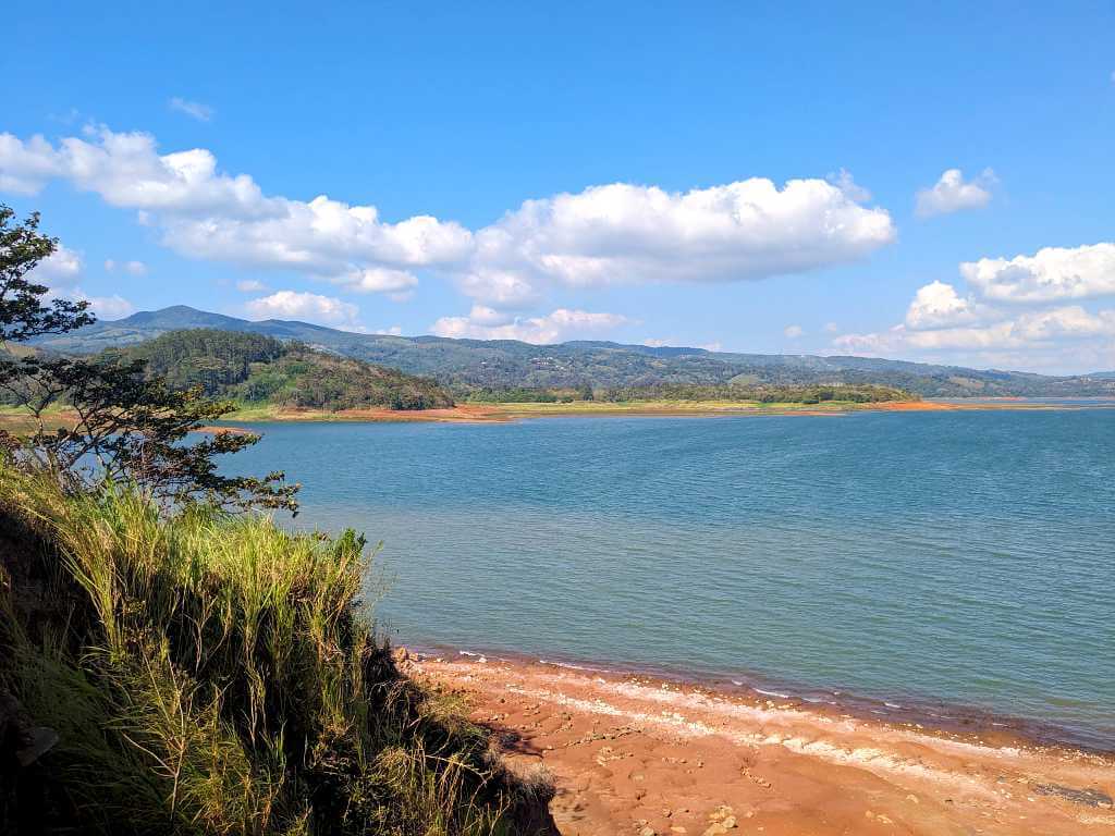 Shores of Arenal Lake on a sunny day with lower water levels, surrounded by green vegetation and distant mountains.