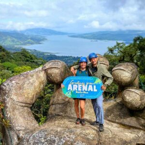 Two travelers posing on La Mano del Arenal, surrounded by lush rainforest with Arenal Lake in the background.