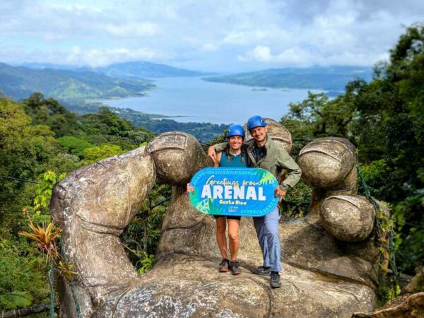 Two travelers posing on La Mano del Arenal, surrounded by lush rainforest with Arenal Lake in the background.