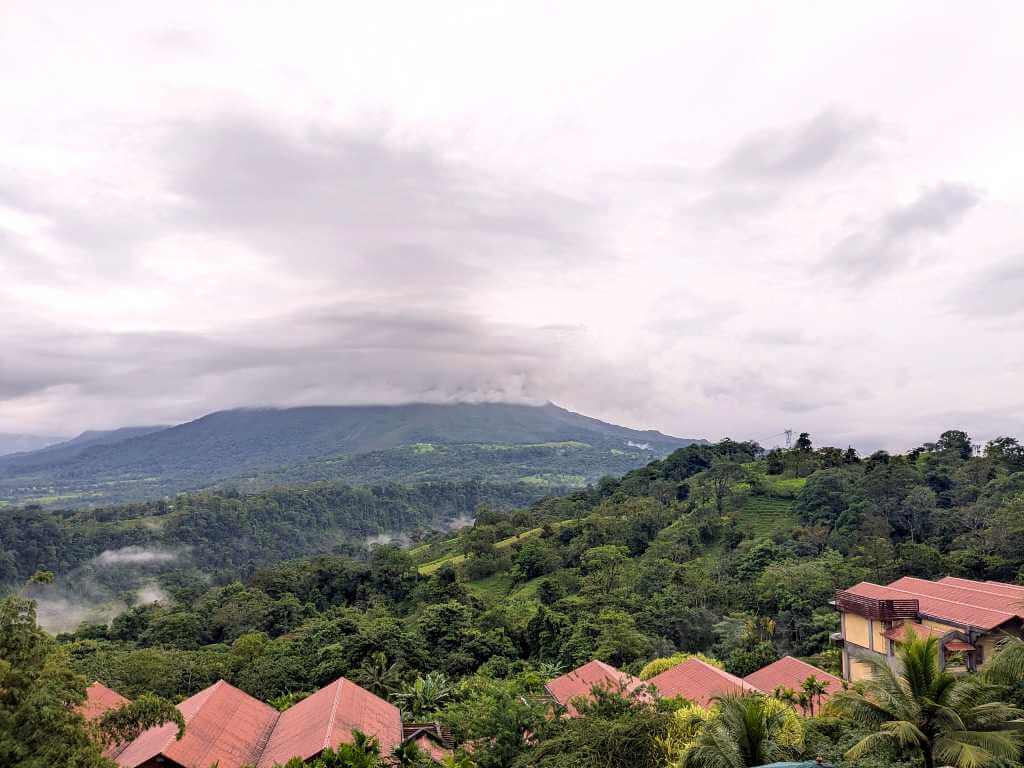 Arenal Volcano partially covered by clouds, rainforest in the middle ground, and a hotel with palm trees in the foreground.