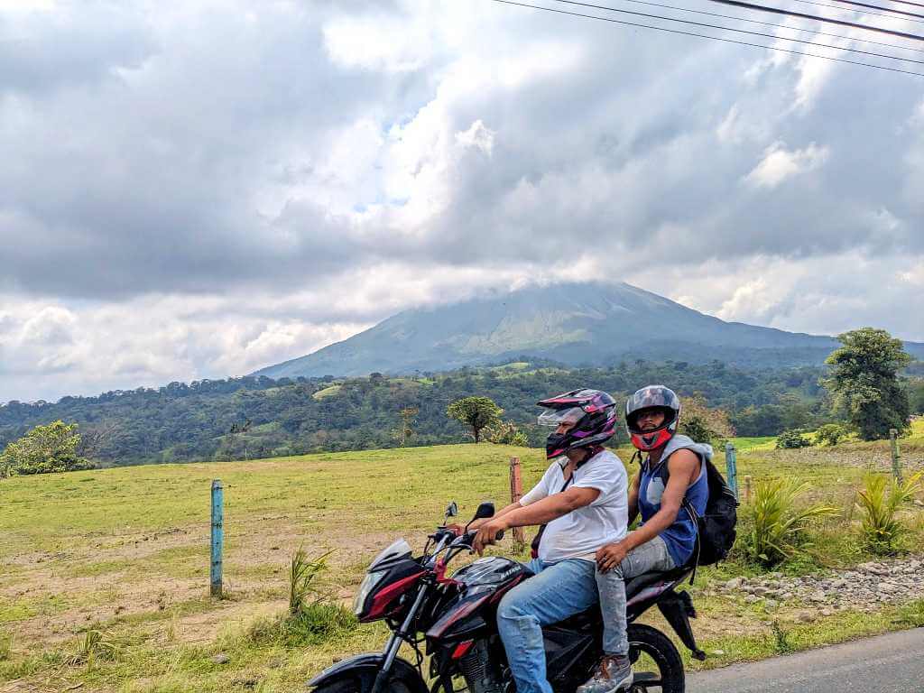 Two Costa Ricans driving a motorcycle in La Fortuna with Arenal Volcano partially covered by clouds in the background.