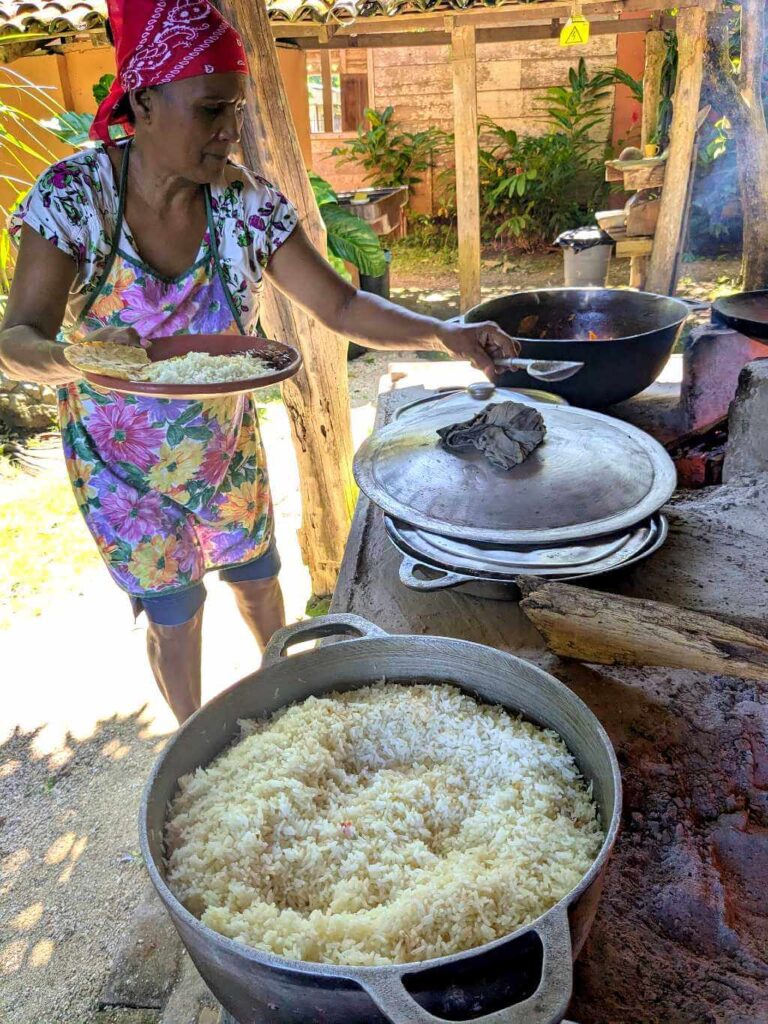 A woman serving lunch, with pots of freshly cooked rice, beans, and other ingredients, all made over a wood fire (fogón), showcasing the clean and fresh preparation process.