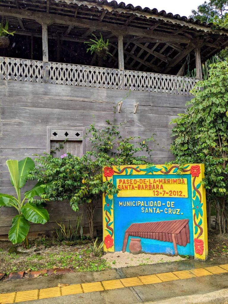 Side perspective of the Casona Cultural sign, local plants, banana tree, and a wooden structure with intricate Guanacaste architectural details.